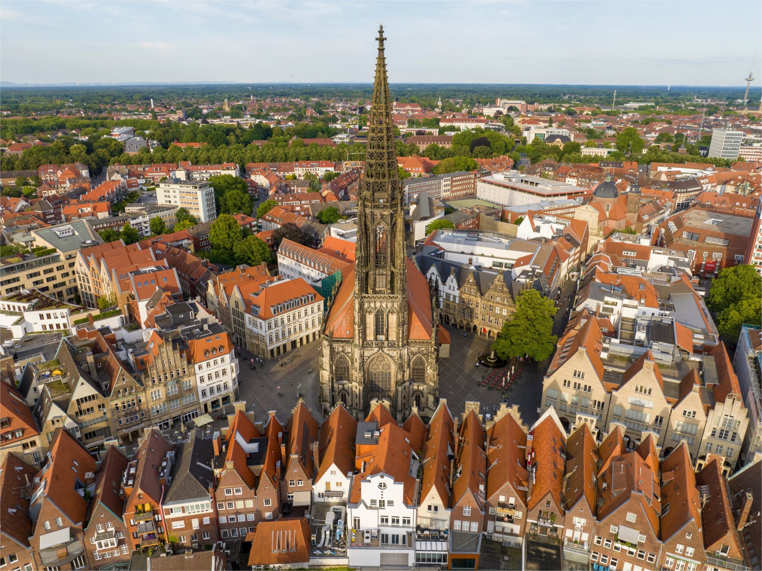 Luftaufnahme von Münster mit dem historischen St.-Paulus-Dom im Zentrum, umgeben von traditionellen roten Ziegeldächern und modernen Gebäuden. Die Stadtlandschaft zeigt eine Mischung aus Altstadtarchitektur und grünen Parkanlagen.