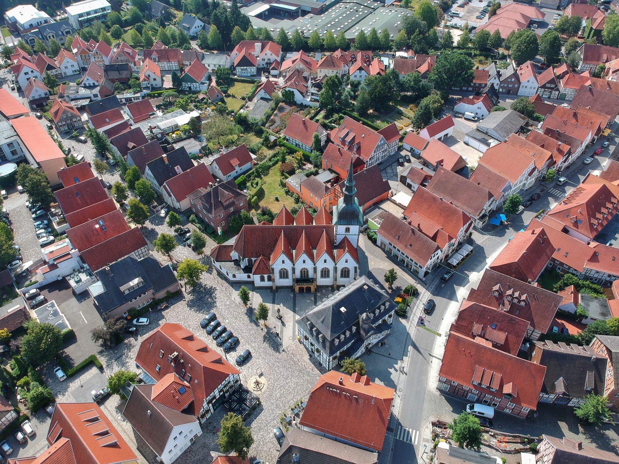 Luftaufnahme einer charmanten Stadt mit roten Dächern, historischen Gebäuden und einem zentralen Platz, umgeben von Wohn- und Geschäftshäusern. Die Aufnahme zeigt die enge Bebauung und die grüne Umgebung, die das Stadtbild prägen.