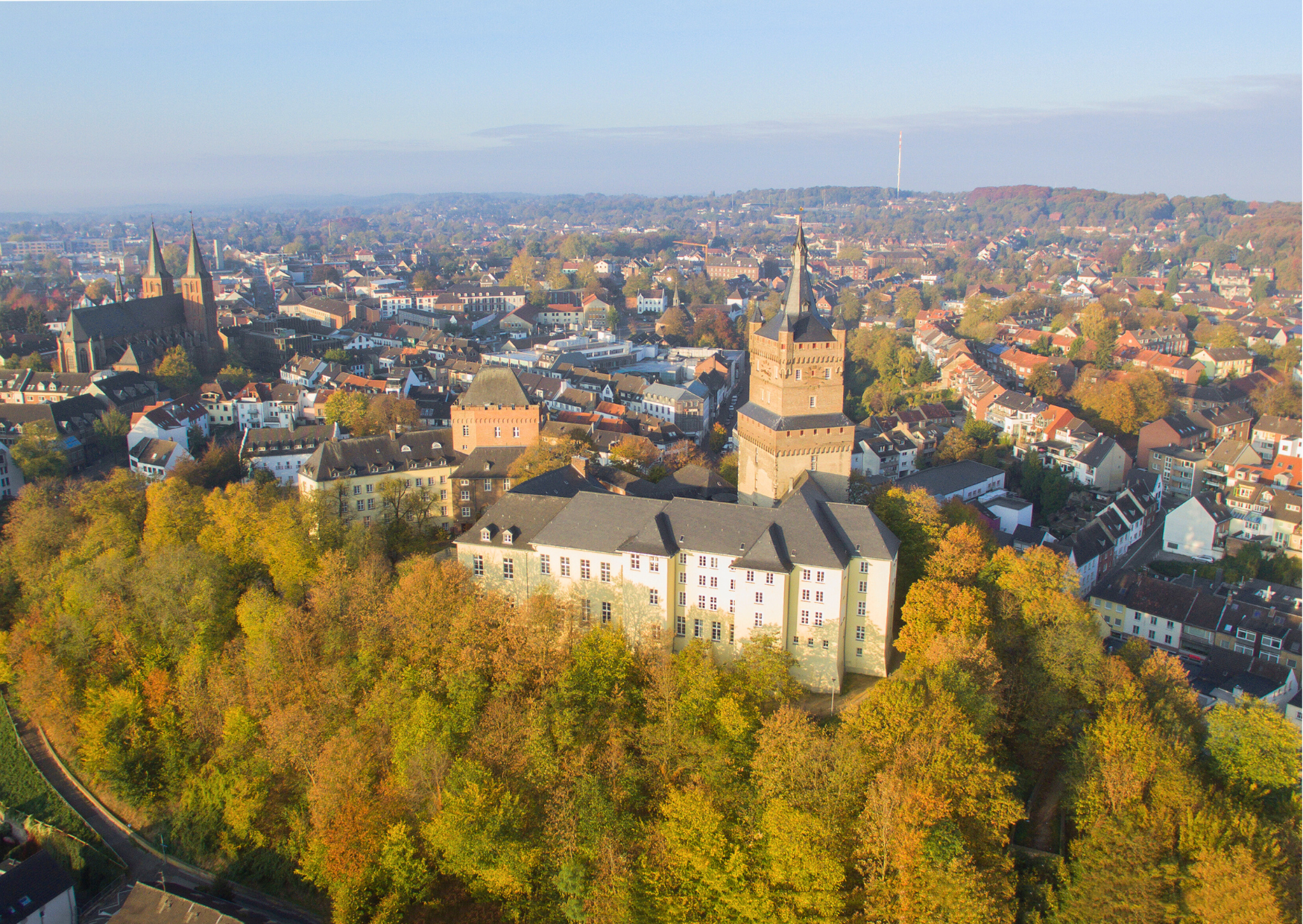 Luftaufnahme einer Stadt mit herbstlichen Bäumen, historischen Gebäuden und einer markanten Burg im Vordergrund, umgeben von einer malerischen Landschaft.