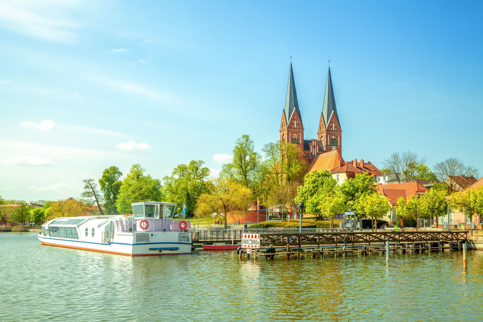 Schöne Aussicht auf ein Boot im Wasser vor einer malerischen Stadt mit zwei markanten Kirchtürmen und grünen Bäumen unter klarem Himmel.